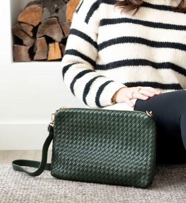 A green woven bag is positioned next to a woman wearing leggings and white/black horizontal striped sweater sitting on the floor.